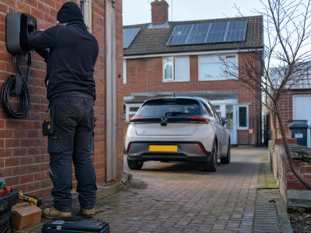 Electrician fitting an EV charger to a brick wall beside a driveway in Birmingham, with solar panels on the roof
